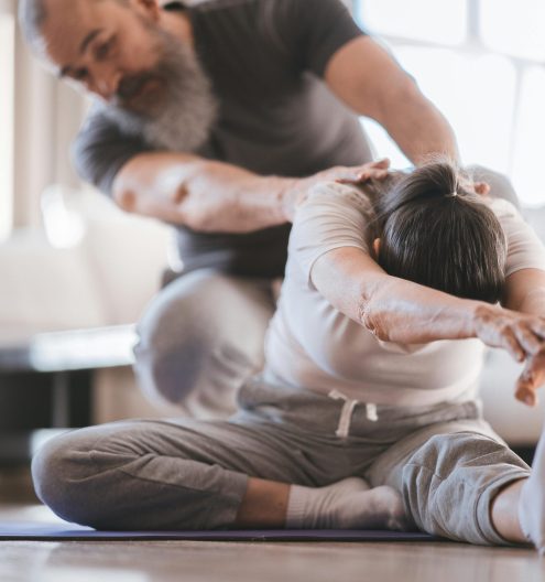 Senior couple practicing yoga at home with assistance, focusing on flexibility and wellness.